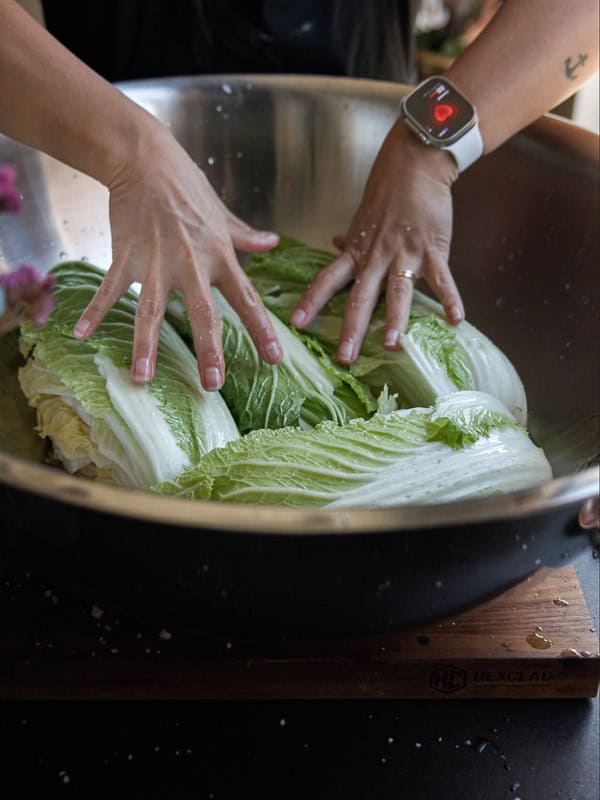 A person wearing a smartwatch is preparing kimchi. They are massaging large cabbage leaves in a metal bowl on a wooden cutting board.